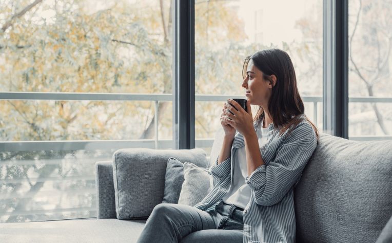 A woman sits on a couch next to floor-to-ceiling windows with sunshine coming through them and holds a mug close to her chin with both hands.
