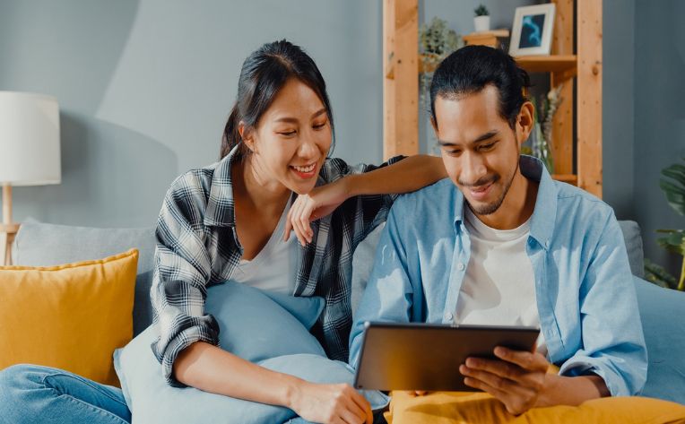 A smiling woman and man sit on a couch with pillows on their laps and look at something on a tablet the man is holding.