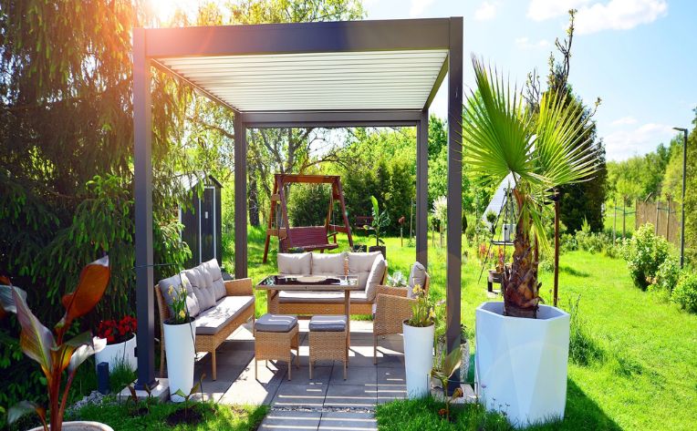 A standalone louvered roof aluminum pergola in a backyard with a stone floor, outdoor furniture under it, a wooden swing and a shed behind it, and an expansive grass yard to the side of it. 