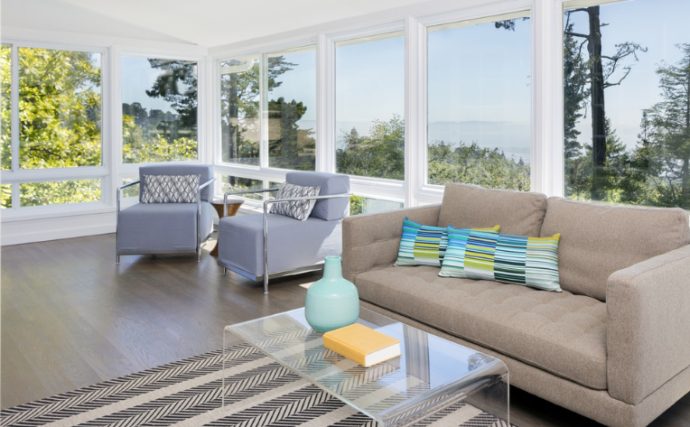 Bright sunroom with large windows, gray seating, a beige sofa, and a glass coffee table overlooking a wooded landscape.