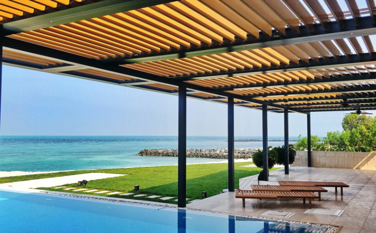 Modern oceanfront patio with a sleek black pergola featuring wood slats overhead, overlooking a turquoise sea. A swimming pool sits in the foreground, with lounge benches on a tiled terrace beside a green lawn and rocky shoreline under a clear blue sky.