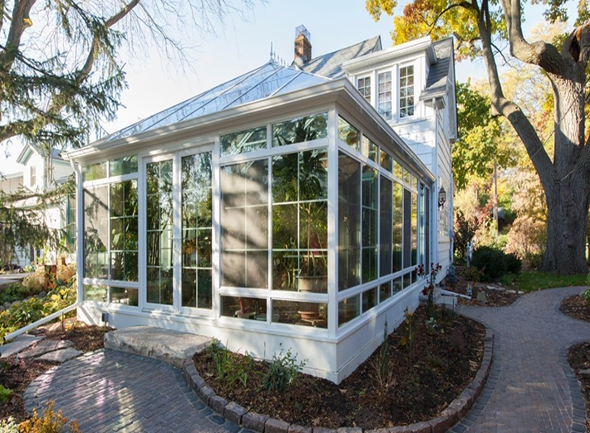 The exterior of a sunroom with white framing and a solid roof attached to a two-story house.