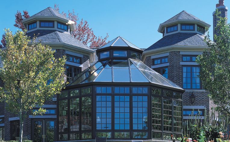 A peaked roof sunroom with all-glass walls and roof attached to a brick house with a garden and patio next to it and a large lawn beyond that. 
