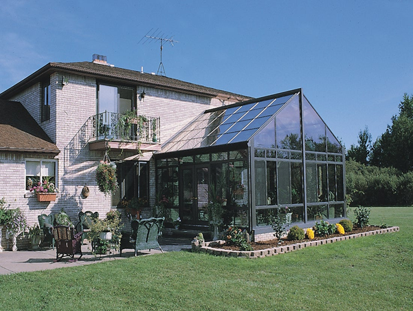 A peaked roof sunroom with all-glass walls and roof attached to a brick house with a garden and patio next to it and a large lawn beyond that.