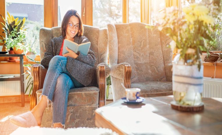 A smiling woman sits in an armchair looking at a book with an empty armchair next to her and a coffee table with a teacup on it in the foreground and sunlight streaming in through the tall windows behind her. 