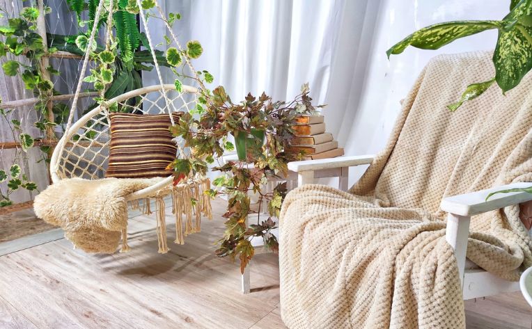 A hanging swing with rope sides suspended over a round jute rug with a small white circular table next to it with a plant on it and the patio room windows in the background.