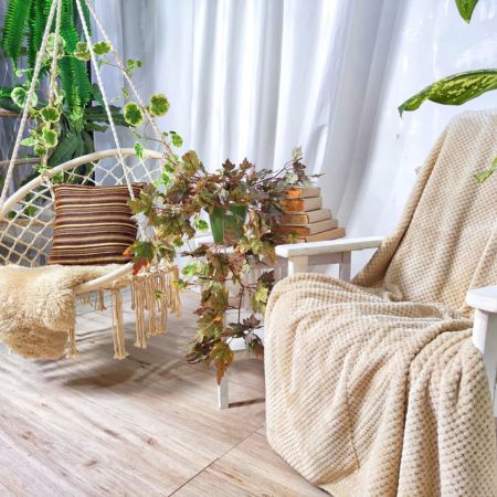 A room with a wicker swing chair with a blanket and pillow on it and a wooden chair with a blanket on it and a table between them with a vine plant on it.