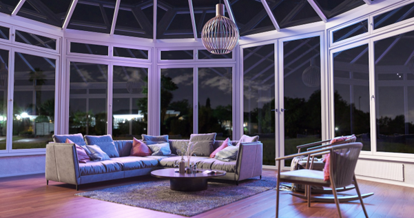 A solarium sunroom with surround glass walls and a glass ceiling with a modern light fixture suspended from the ceiling and an L-shaped couch, area rug, and round coffee table in the middle. 